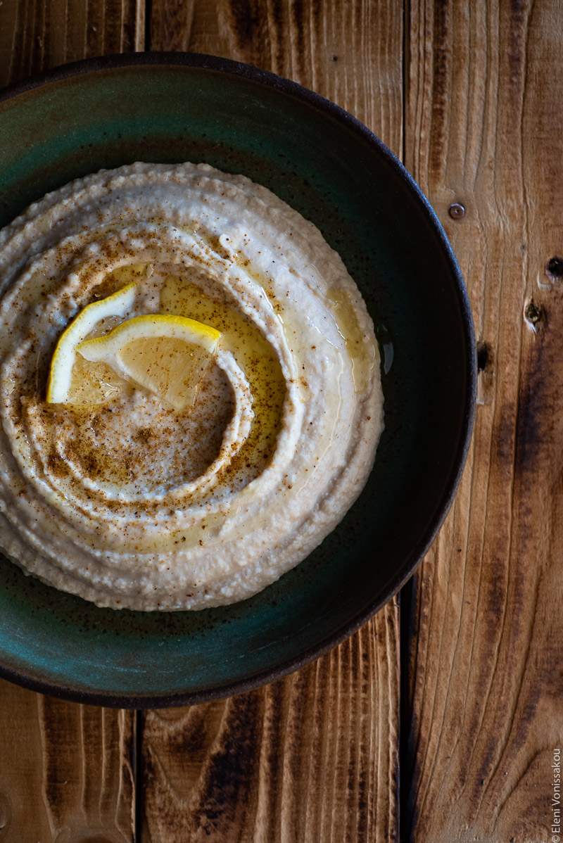 Hummus-Style White Bean Dip with Ras El Hanout www.thefoodiecorner.gr Photo description: A close view of a ceramic green dish (slightly off centre) with humms on a distressed wooden background. The hummus is dusted with ras el hanout, drizzled with olive oil and garnished with a couple of lemon wedges.