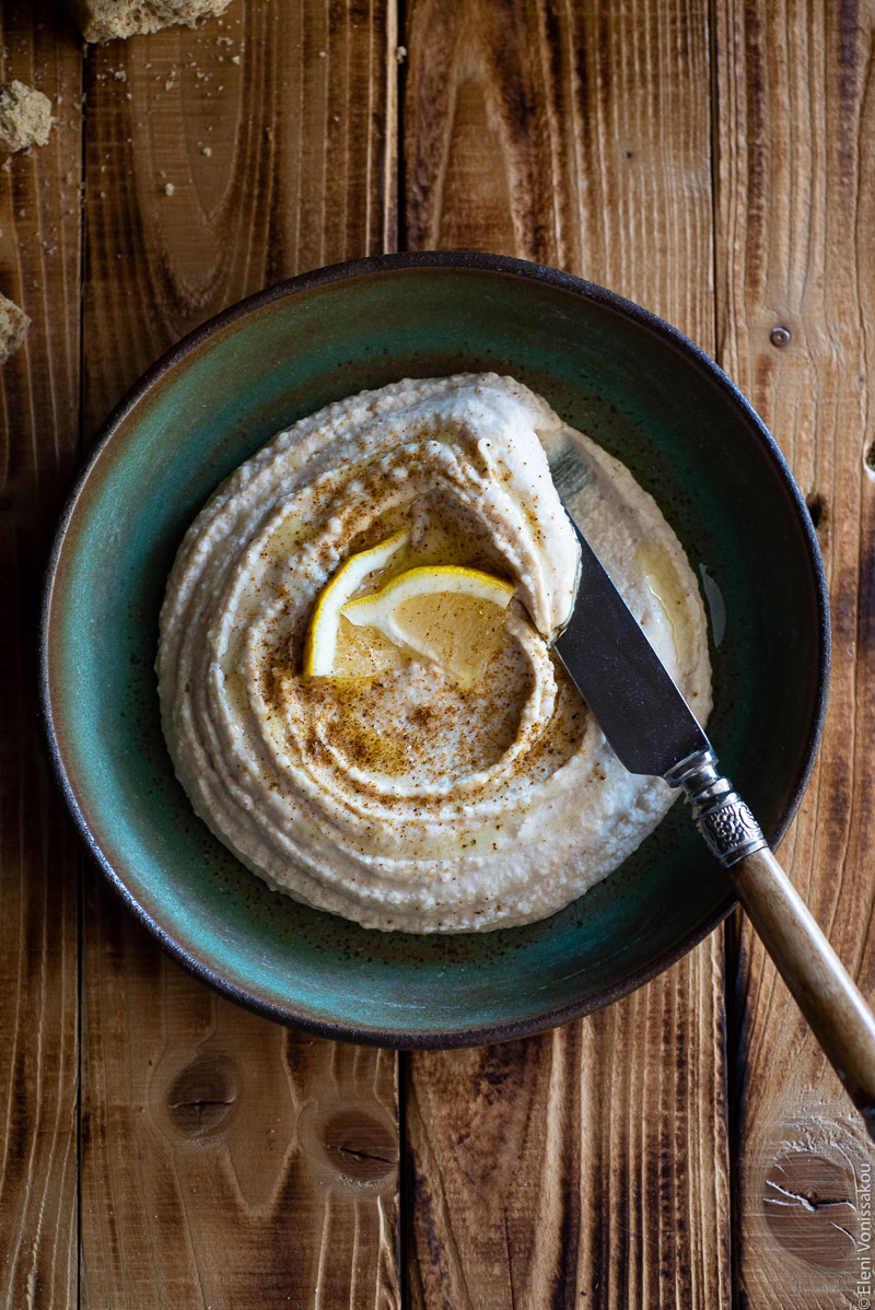 Hummus-Style White Bean Dip with Ras El Hanout www.thefoodiecorner.gr Photo description: A ceramic green dish with humms on a distressed wooden background. The hummus is dusted with ras el hanout, drizzled with olive oil and garnished with a couple of lemon wedges. Resting on the side of the dish is a knife that has been dipped into the hummus.