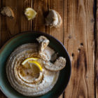 Hummus-Style White Bean Dip with Ras El Hanout www.thefoodiecorner.gr Photo description: A ceramic green dish with humms on a distressed wooden background. The hummus is dusted with ras el hanout, drizzled with olive oil and garnished with a couple of lemon wedges. At the side are two broken rusks half dipped into the hummus. To the top of the image directly on the wood are some more broken rusks and two lemon wedges, one of them squeezed.