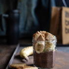 Chocolate Chia Pudding www.thefoodiecorner.gr Photo description: A jar of chocolate chia pudding with slices of banana and pieces of biscuit on top. The jar is on a rusty coloured surface and next to it are a biscuit and a banana. In the background is a lunch bag, a jeans jacket and a travel mug.