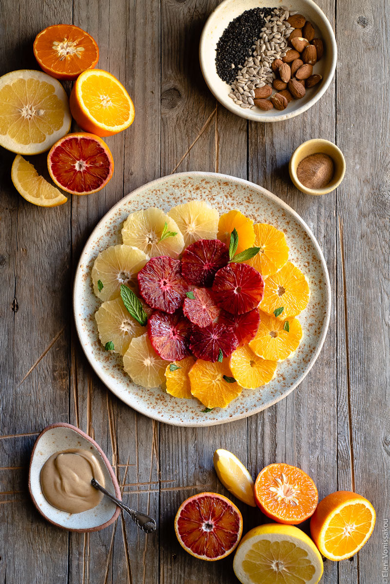 Acerola and Coconut Yoghurt Citrus Fruit Platter www.thefoodiecorner.gr Photo description: A large ceramic platter with slices of citrus fruit arranged in concentric circles. To the top right of the photo is a small dish with nuts and seeds. To the bottom left is a small plate with peanut butter and a spoon. On the top left and bottom right are halved citrus fruits.