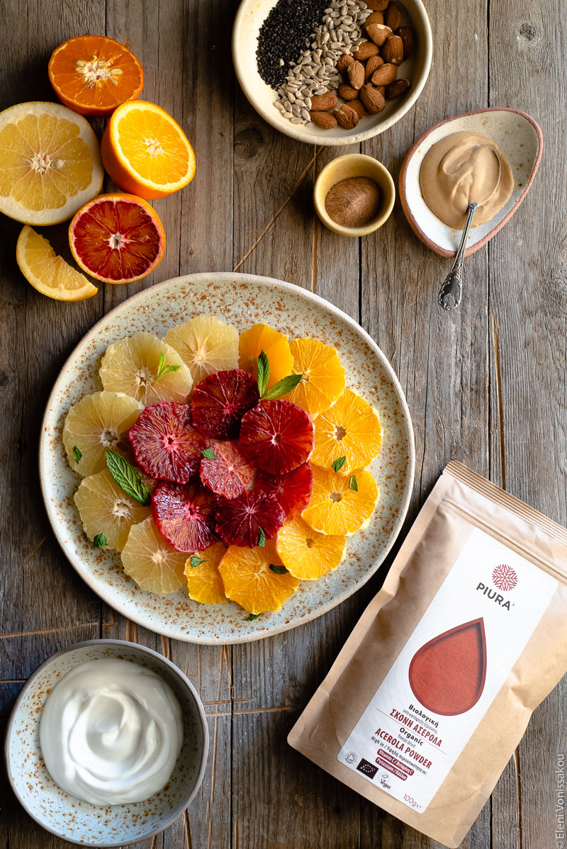 Acerola and Coconut Yoghurt Citrus Fruit Platter www.thefoodiecorner.gr Photo description: A large ceramic platter with slices of citrus fruit arranged in concentric circles. To the top right of the photo is a small dish with nuts and seeds and a plate with peanut butter. To the bottom left is bowl of coconut yoghurt. On the top left are some halved citrus fruits and on the bottom right is a packet of acerola.