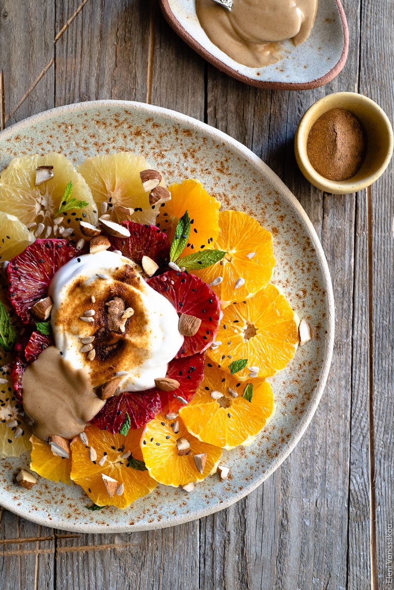 Acerola and Coconut Yoghurt Citrus Fruit Platter www.thefoodiecorner.gr Photo description: A close up of a large ceramic platter with slices of citrus fruit arranged in concentric circles. On top of the fruit is a dollop of coconut yoghurt, peanut butter and a sprinkling of nuts and seeds. To the top right of the photo is a small plate with peanut butter and a tiny bowl of acerola powder. 