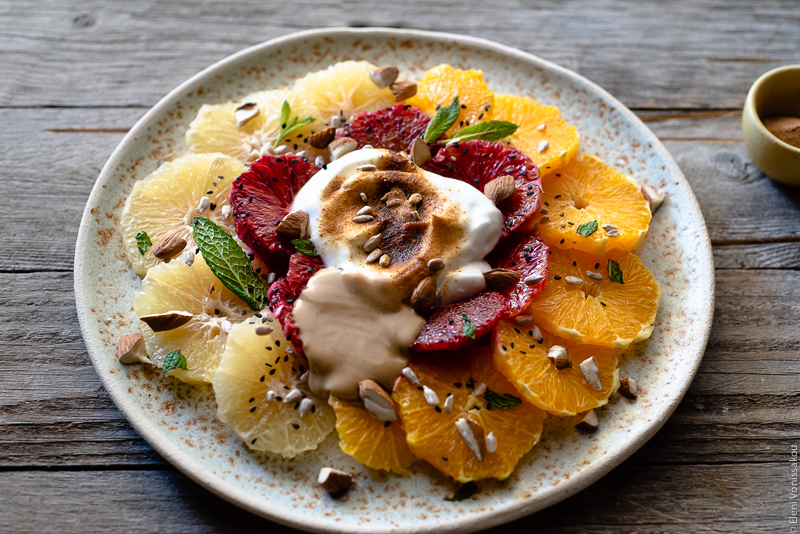 Acerola and Coconut Yoghurt Citrus Fruit Platter www.thefoodiecorner.gr Photo description: A close up side view of a large ceramic platter with slices of citrus fruit arranged in concentric circles. On top of the fruit is a dollop of coconut yoghurt, peanut butter and a sprinkling of nuts and seeds. 