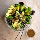 Leafy Green Salad with Blueberries and Savoury Buckwheat Granola www.thefoodiecorner.gr Photo description: A large glass bowl full of salad leaves with slices of cucumber and avocado arranged on top. Some blueberries and granola are also scattered on the salad. A set of serving spoons are in the bowl. To the right of the bowl is another small one with dressing inside. Both are sitting on a pretty marble surface.