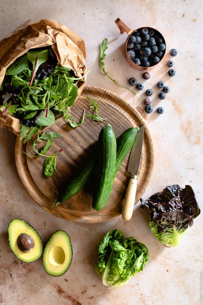 Leafy Green Salad with Blueberries and Savoury Buckwheat Granola www.thefoodiecorner.gr Photo description: A chopping board with small cucumbers on it and a knife. To the top left is a paper bag with salad leaves spilling out. To the top right is a measuring cup with blueberries, a few of them spilled around it. To the bottom right are two baby lettuces and to the bottom left is a halved avocado.