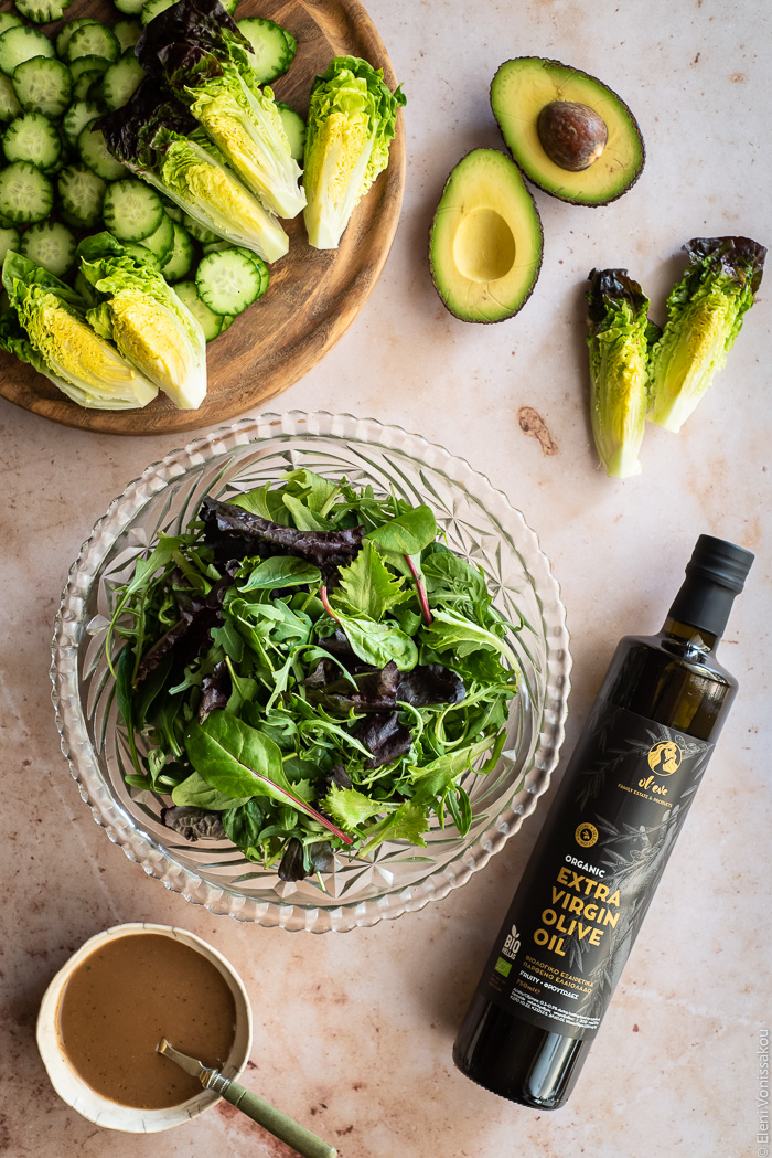 Leafy Green Salad with Blueberries and Savoury Buckwheat Granola www.thefoodiecorner.gr Photo description: A large glass bowl full of salad leaves. To the top left is a chopping board with sliced cucumber and quartered baby lettuces. To the top right is a halved avocado and to the right of the bowl is a bottle of olive oil. To the bottom left of the image is a small ceramic bowl of dressing.