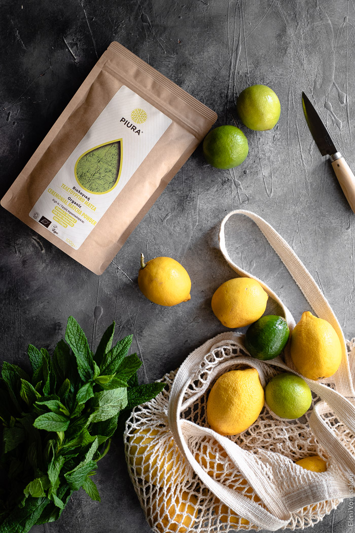 Easy Homemade Matcha Lemonade www.thefoodiecorner.gr Photo description: A string bag with lemons and limes spilling out. To the side of it a bunch of spearmint. To the top of the photo is a packet of Piura matcha and a sharp knife.