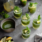 Easy Homemade Matcha Lemonade www.thefoodiecorner.gr Photo description: Six glasses of matcha lemonade on a grey surface. Around them are a glass jar with lemonade concentrate, a small ceramic bowl of matcha powder, a small dish with cut lemons and limes, and a ceramic jug with prepared matcha tea. In the bottom right corner is a bunched up linen tea towel. The matcha lemonade has pretty layers, with the lemonade at the bottom of the glasses and the matcha sitting on top. The drinks are garnished with small wedges of lemons and limes, and spearmint leaves.