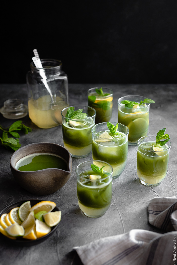 Easy Homemade Matcha Lemonade www.thefoodiecorner.gr Photo description: A 3/4 view of six glasses of matcha lemonade, their two layers visible (lemonade at the bottom and matcha sitting on top). To the side are a dish of cut lemons and limes, a ceramic jug of prepared matcha, some spearmint barely visible and a glass jar of lemonade concentrate in the background.