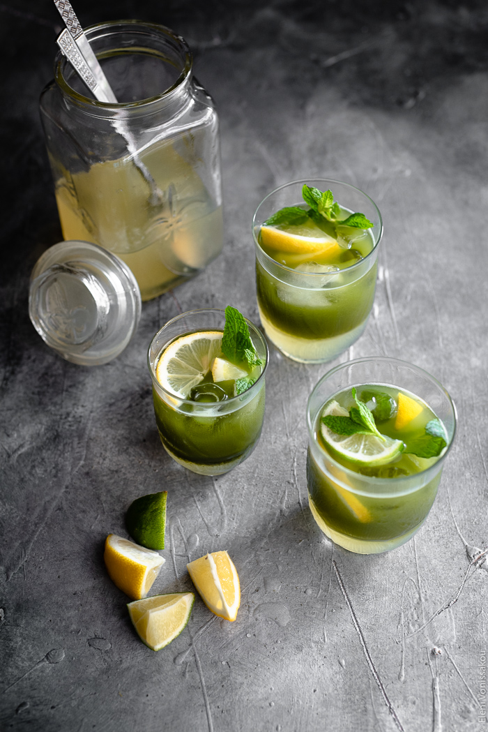 Easy Homemade Matcha Lemonade www.thefoodiecorner.gr Photo description: A 3/4 view of three glasses of matcha lemonade, their two layers visible (lemonade at the bottom and matcha sitting on top). In front of them are some small wedges of lemon and lime. Behind them is a jar of lemonade concentrate.