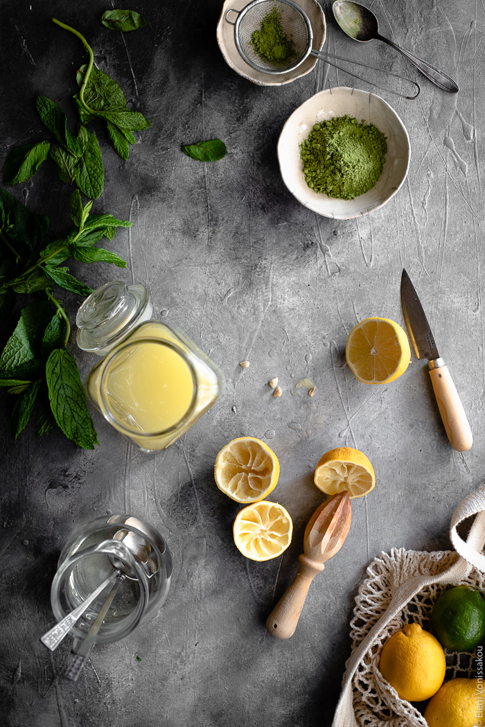 Σπιτική Λεμονάδα με Τσάι Matcha www.thefoodiecorner.gr Photo description: Some juiced lemon halves, a wooden juicer, a knife, two tiny bowls of matcha, a bunch of spearmint, a glass jar of lemonade concentrate and a small jug of simple syrup, all laid out on a grey surface. Barely visible in the bottom corner is a string bag with lemons and limes. 