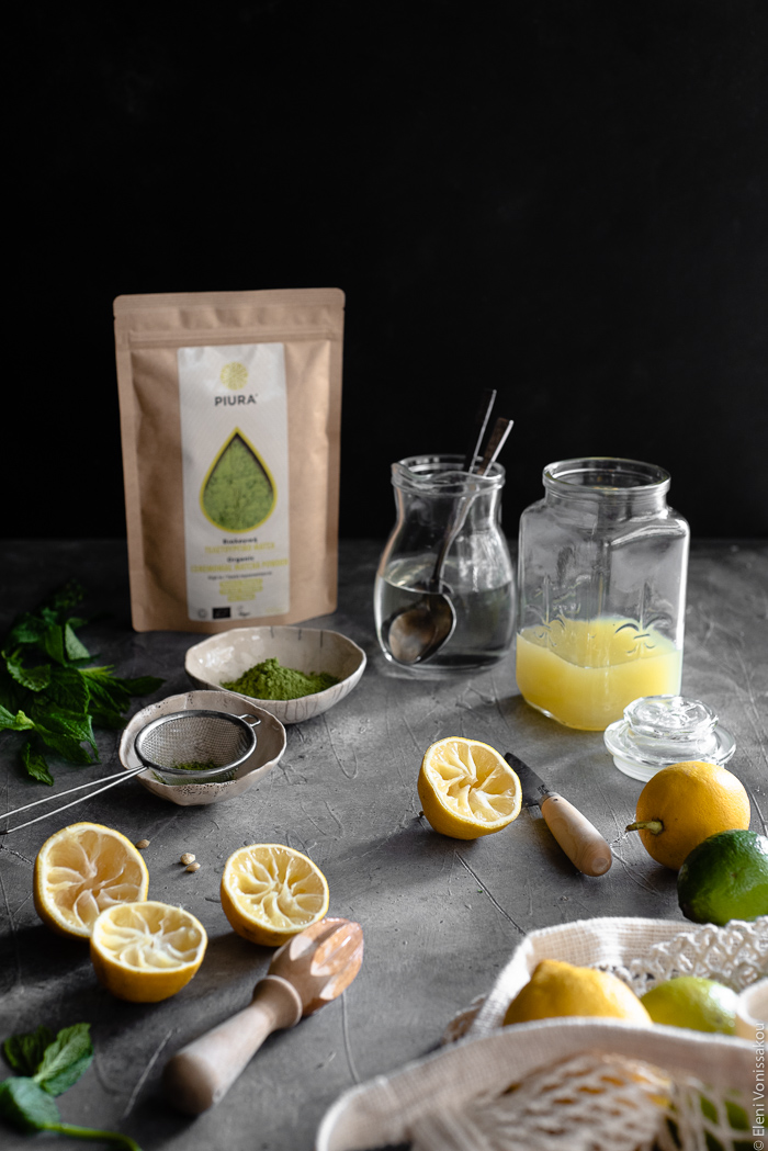 Easy Homemade Matcha Lemonade www.thefoodiecorner.gr Photo description: A side view of a surface with lemonade making items. Squeezed lemon halves, a glass jar with lemonade concentrate, two tiny bowls of matcha powder, a small jug of simple syrup and a packet of Piura matcha in the background.