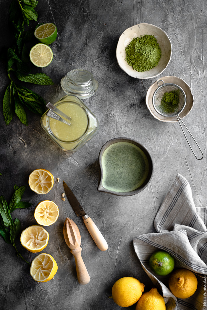 Easy Homemade Matcha Lemonade www.thefoodiecorner.gr Photo description: A top-down view of a surface with lemonade making items. Squeezed lemon halves, a glass jar with lemonade concentrate, two tiny bowls of matcha powder, a small jug of simple syrup, a knife and wooden juicer, and a ceramic bowl of prepared matcha tea. In the bottom corner are some lemons and limes.