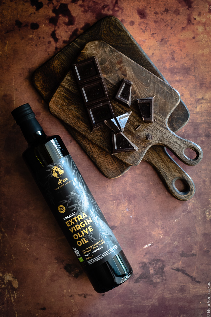 Easy, Egg-Free Olive Oil Chocolate Mousse www.thefoodiecorner.gr Photo description: A bottle of olive oil lying on a rusty coloured surface next to a chopping board with pieces of dark chocolate on it.