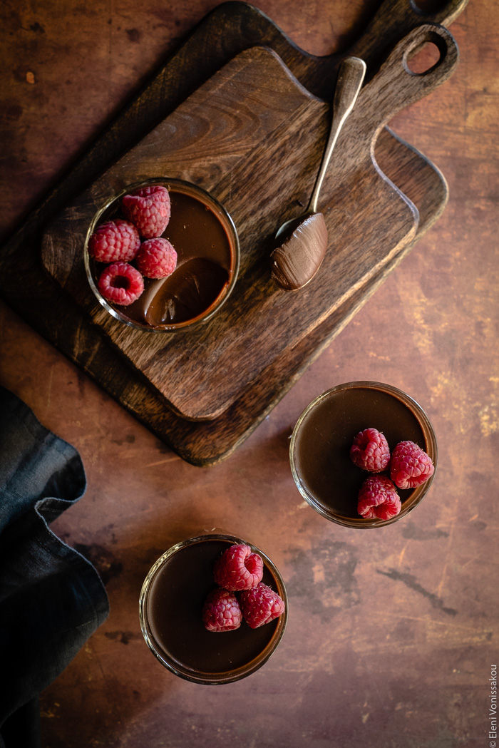 Easy, Egg-Free Olive Oil Chocolate Mousse www.thefoodiecorner.gr Photo description: A top down view of three glasses of chocolate mousse with raspberries on top. The glass at the back is sitting on two stacked chopping boards. A spoon with some mousse on is lying next to the glass on the board.