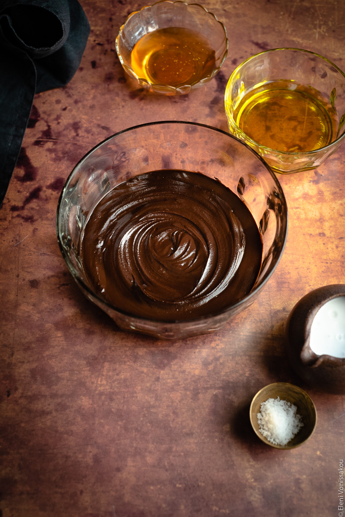 Easy, Egg-Free Olive Oil Chocolate Mousse www.thefoodiecorner.gr Photo description: Top view of a large glass bowl with melted chocolate inside. To the right are small bowls of oil and honey, a ceramic jug with coconut milk and a tiny brass bowl of salt.