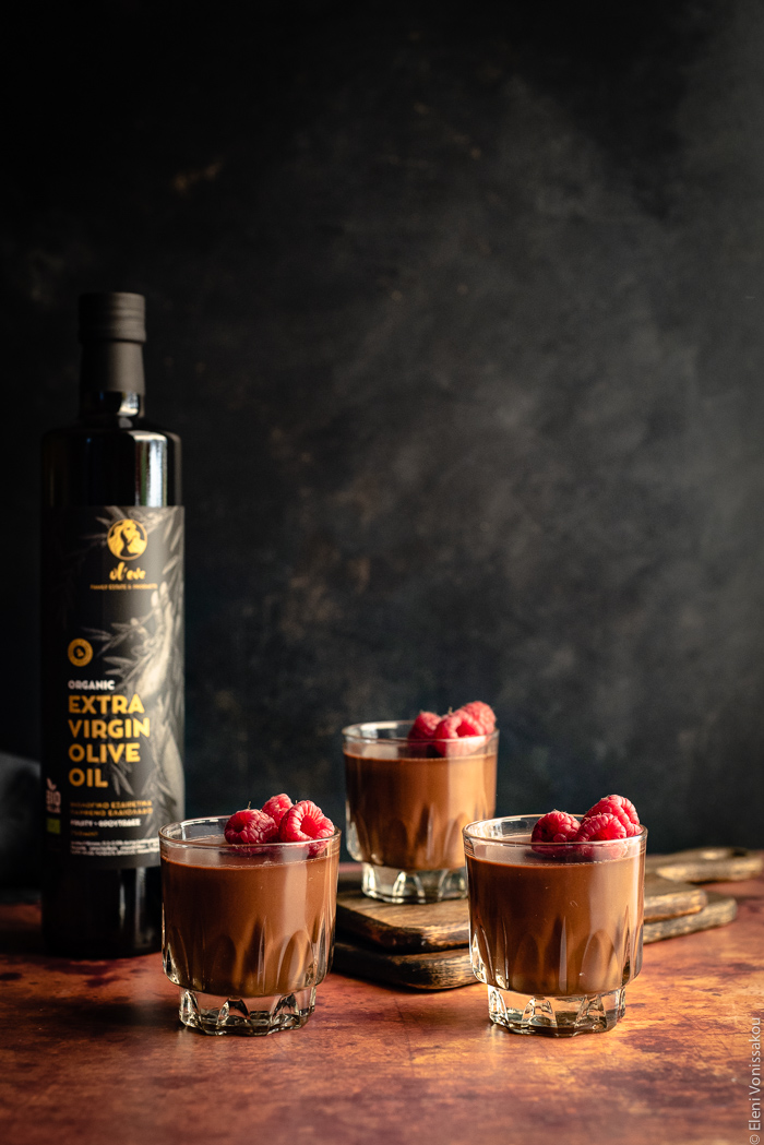 Easy, Egg-Free Olive Oil Chocolate Mousse www.thefoodiecorner.gr Photo description: Side view of three glasses of chocolate mousse against a dark background. The mousse is garnished with raspberries. The glass furthest away is sitting on two wooden chopping boards, thus it is slightly higher than the front two. To the left is a bottle of olive oil.