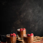 Easy, Egg-Free Olive Oil Chocolate Mousse www.thefoodiecorner.gr Photo description: Side view of three glasses of chocolate mousse against a dark background. The mousse is garnished with raspberries. The glass furthest away is sitting on two wooden chopping boards, thus it is slightly higher than the front two.