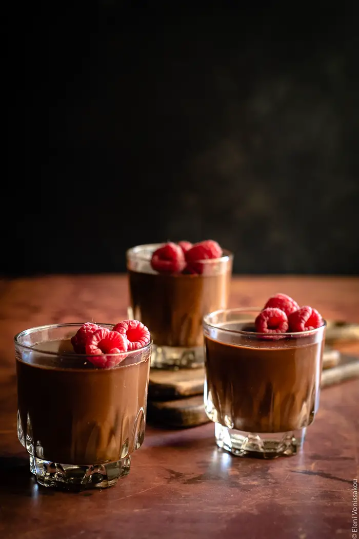 Easy, Egg-Free Olive Oil Chocolate Mousse www.thefoodiecorner.gr Photo descriptions: A closer side view of three pretty glasses of chocolate mousse with raspberries on top. The glass at the back is sitting on two stacked chopping boards.