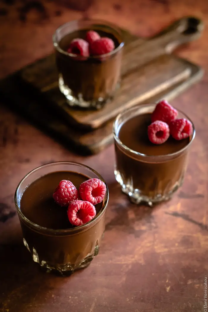Easy, Egg-Free Olive Oil Chocolate Mousse www.thefoodiecorner.gr Photo description: A ¾ view of three pretty glasses of chocolate mousse with raspberries on top. The glass at the back is sitting on two stacked chopping boards.