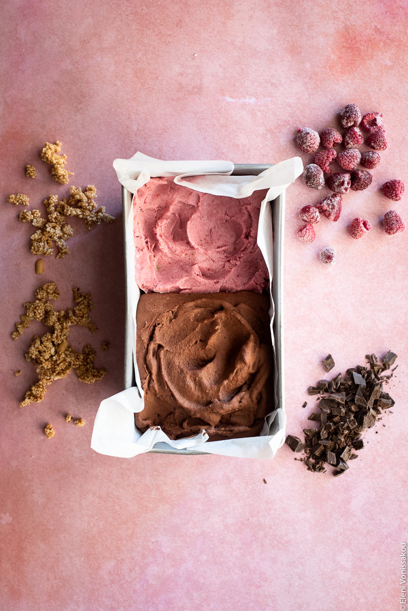 Four Ingredient Banana Ice Cream - Raspberry or Chocolate www.thefoodiecorner.gr Photo description: A baking tin lined with baking paper, filled on one side with chocolate ice cream and on the other with raspberry. To the left of the tin sprinkled on the pink surface is some chopped sesame granola bar and to the right are some chopped dark chocolate and some frozen raspberries.