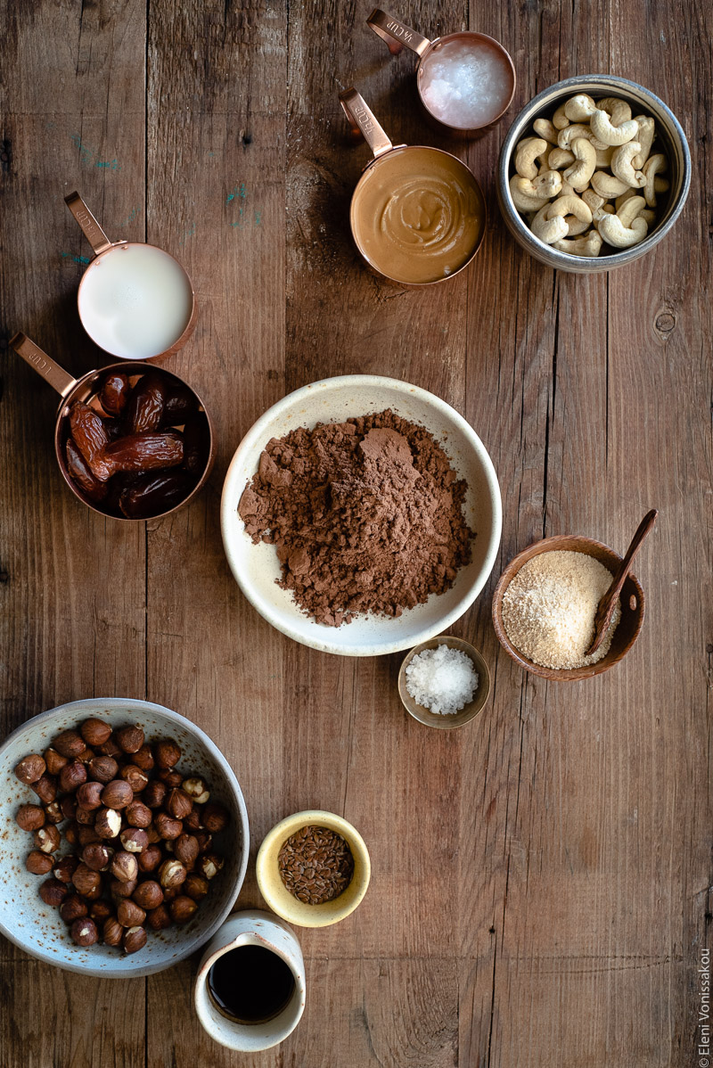 No Bake Cacao Peanut Butter Tart with a Hazelnut Base (Plant-Based) www.thefoodiecorner.gr Photo description: Ingredients arranged on a wooden surface. At the top left are two measuring cups with oat milk and dates. In the top right corner is a measuring cup with coconut oil, another with peanut butter and a ceramic cup with cashews. In the centre of the image is a bowl of cacao powder, and beside it are a small bowl of maple sugar and a tiny brass bowl of salt flakes. In the bottom left corner is a ceramic bowl of hazelnuts, a tiny bowl of flax seeds and a tiny jug of coffee.