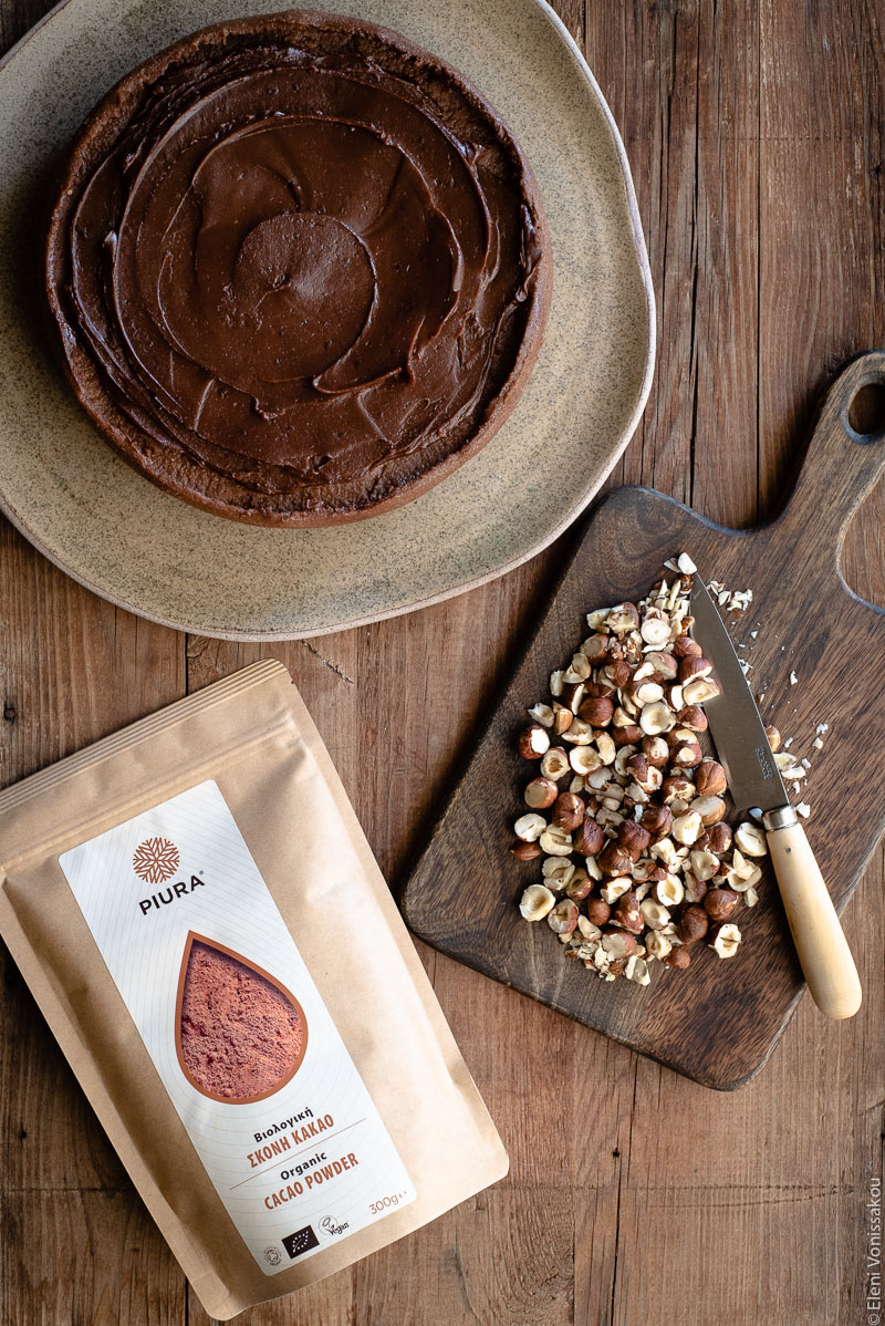 No Bake Cacao Peanut Butter Tart with a Hazelnut Base (Plant-Based) www.thefoodiecorner.gr Photo description: A chocolate covered tart on a large ceramic plate in the top left corner of the image. On the right is a chopping board with hazelnuts on it and a knife. In the bottom left corner is a packet of cacao powder.