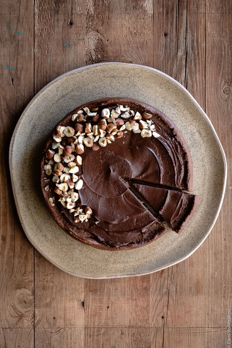 No Bake Cacao Peanut Butter Tart with a Hazelnut Base (Plant-Based) www.thefoodiecorner.gr Photo description: A chocolate covered tart on a large ceramic plate in the centre of the image. The tart is decorated on the left side with chopped hazelnuts. On the right side a slice has been cut and slightly pulled out.