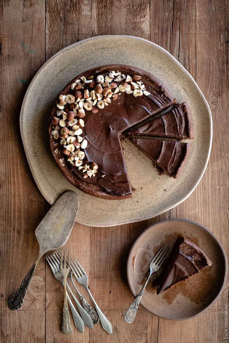 No Bake Cacao Peanut Butter Tart with a Hazelnut Base (Plant-Based) www.thefoodiecorner.gr Photo description: A chocolate covered tart on a large ceramic plate. To the bottom is a small plate with a slice of the tart on it along with a small fork. To the left of the small plate are some more forks and a cake slice lying upside-down.