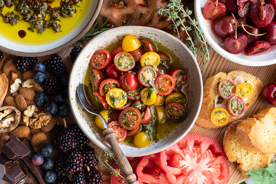 Bruschetta Board with Pickled Cherries, Marinated Cherry Tomatoes, Apricot Salsa and Olive Oil Dip www.thefoodiecorner.gr Photo description: A close up of a bowl of marinated cherry tomatoes with herbs. A wooden spoon is laying in the bowl to the side. There are red, black and yellow tomatoes.