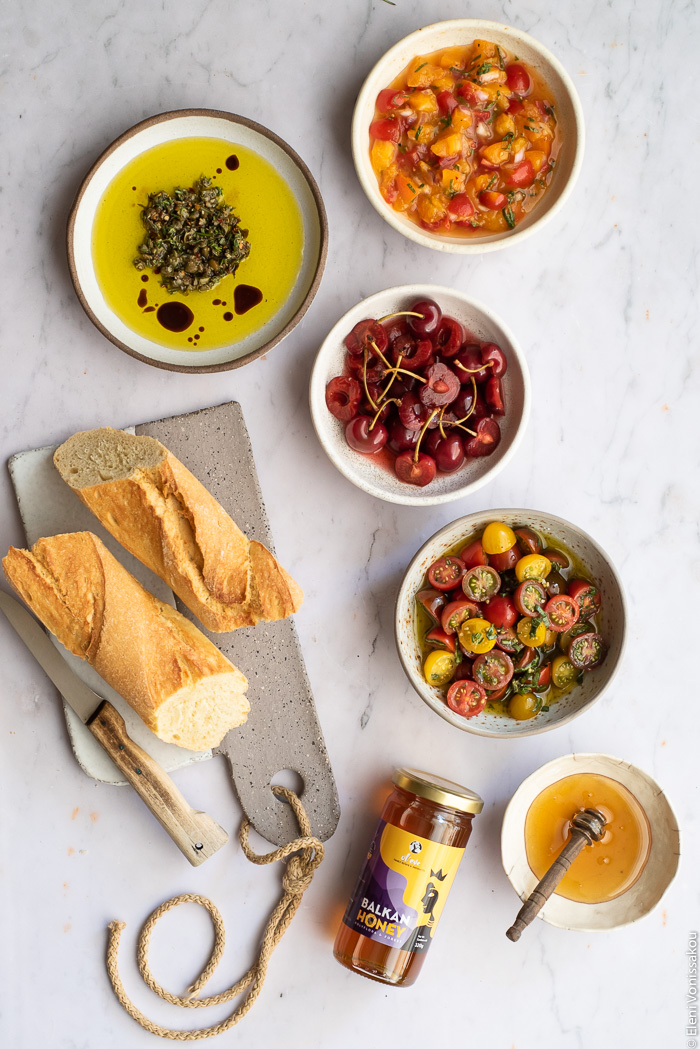 Bruschetta Board with Pickled Cherries, Marinated Cherry Tomatoes, Apricot Salsa and Olive Oil Dip www.thefoodiecorner.gr Photo description: Four ceramic bowls each with a bruschetta topping, one with pickled cherries, one with marinated tomatoes, one with apricot salsa and one will olive oil dip. Beside them a ceramic chopping board with a knife and some baguette bread. To the bottom a smaller bowl and a jar of honey. 