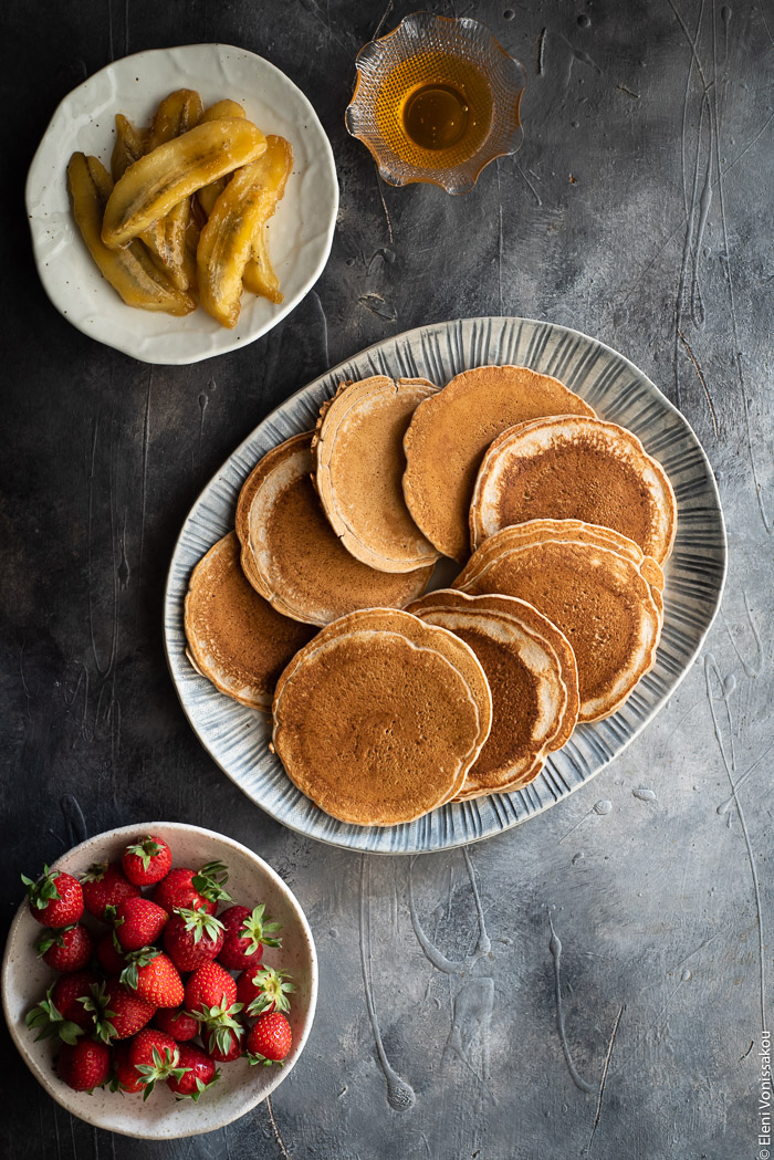 Spelt Pancakes with Honey Fried Bananas (egg and dairy free) www.thefoodiecorner.gr Photo description: A ceramic platter full of pancakes. To the bottom of the platter is a small plate of strawberries and to the top is a plate of fried bananas and a small bowl of honey.