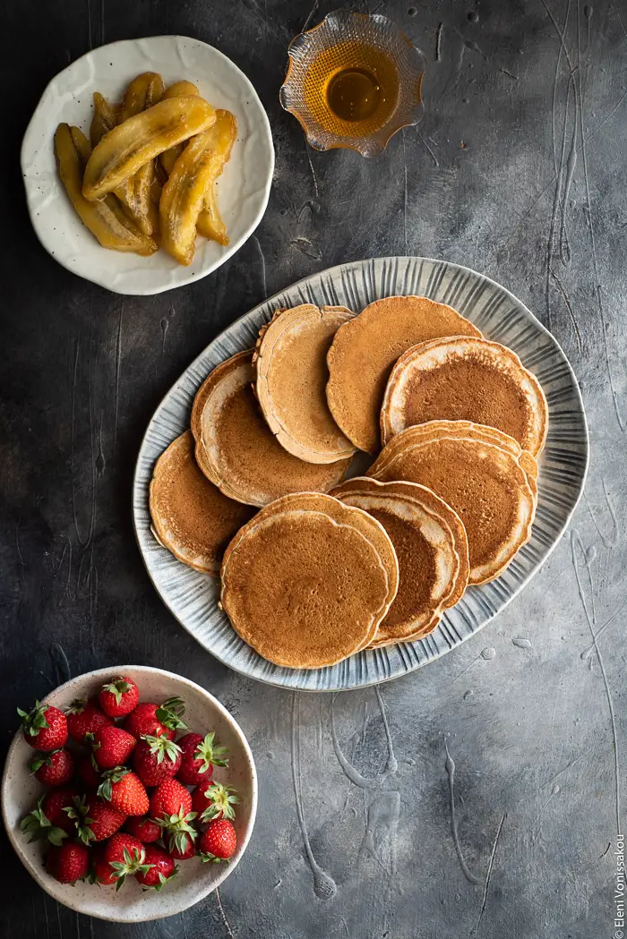 Pancakes με Αλεύρι Σπέλτ και Μελωμένες Τηγανιτές Μπανάνες (χωρίς αυγά) www.thefoodiecorner.gr Photo description: A ceramic platter full of pancakes. To the bottom of the platter is a small plate of strawberries and to the top is a plate of fried bananas and a small bowl of honey.