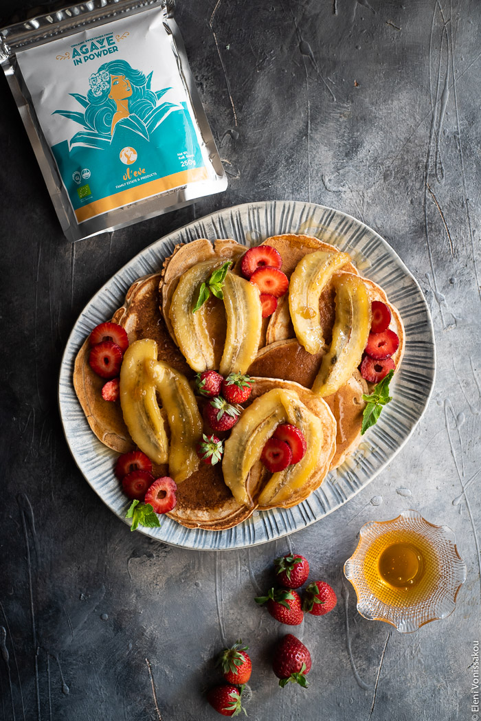 Spelt Pancakes with Honey Fried Bananas (egg and dairy free) www.thefoodiecorner.gr Photo description: A ceramic platter full of pancakes garnished with honey fried bananas and strawberries. To the bottom of the platter are some strawberries and a small bowl of honey. To the top is a packet of agave sugar.