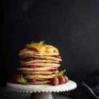 Pancakes με Αλεύρι Σπέλτ και Μελωμένες Τηγανιτές Μπανάνες (χωρίς αυγά) www.thefoodiecorner.gr Photo description: A stack of spelt pancakes on a small ceramic cake stand garnished with honey fried bananas on top and strawberries all around. There is honey dripping down the side of the pancake stack.