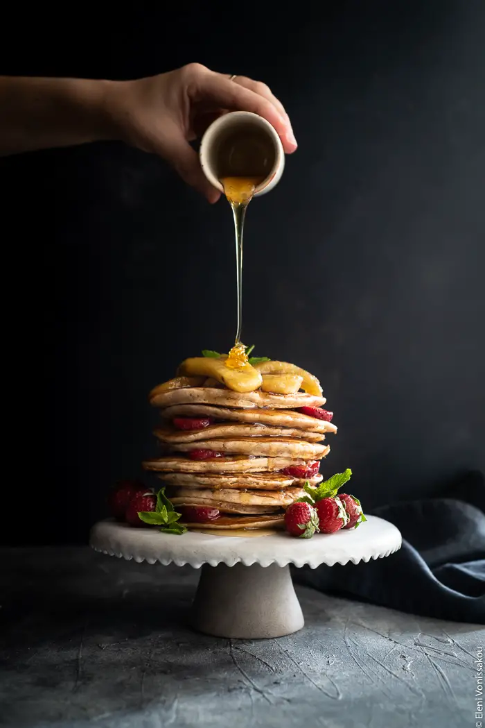 Spelt Pancakes with Honey Fried Bananas (egg and dairy free) www.thefoodiecorner.gr Photo description: A stack of spelt pancakes on a small ceramic cake stand garnished with honey fried bananas on top and strawberries all around. A hand is pouring honey over the top of the stack and it is dripping down the side of the pancakes.