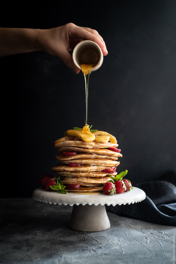 Pancakes με Αλεύρι Σπέλτ και Μελωμένες Τηγανιτές Μπανάνες (χωρίς αυγά) www.thefoodiecorner.gr Photo description: A stack of spelt pancakes on a small ceramic cake stand garnished with honey fried bananas on top and strawberries all around. A hand is pouring honey over the top of the stack and it is dripping down the side of the pancakes.