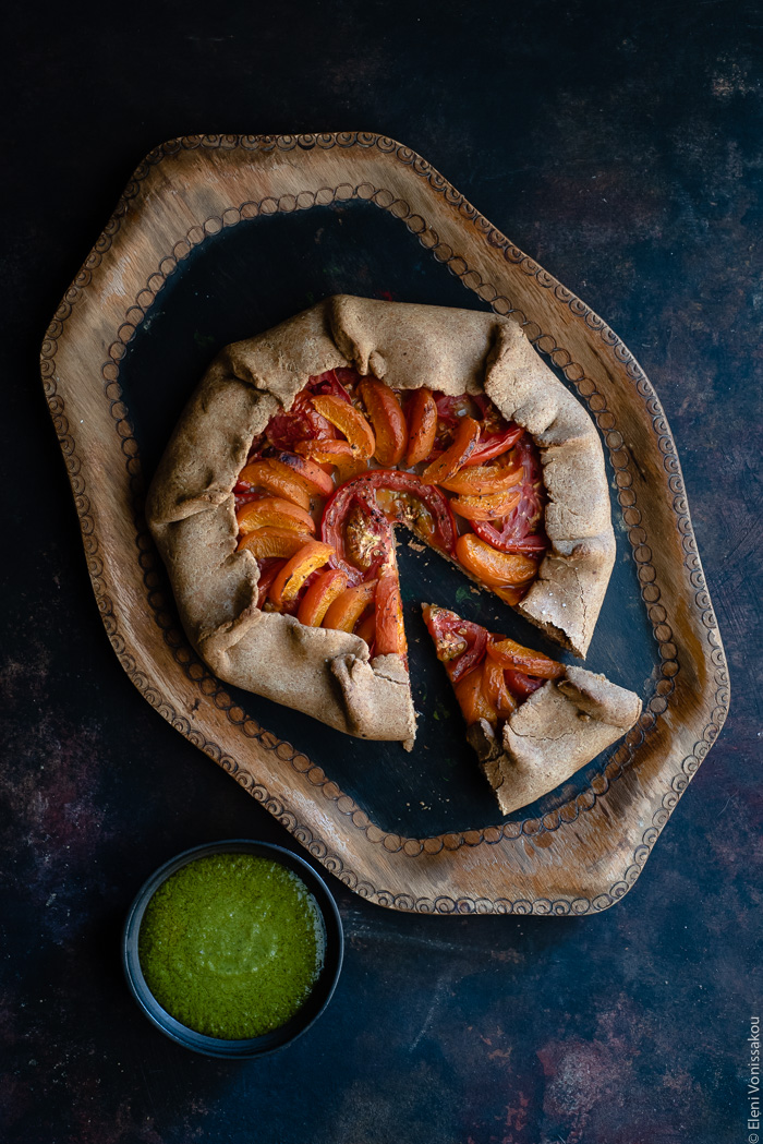 Apricot and Tomato Spelt Galette with Basil Vinaigrette www.thefoodiecorner.gr Photo description: A galette with apricot and tomato slices arranged nicely inside. The galette is on a wooden tray and one slice has been cut and pulled out slightly. At the bottom left of the photo is a small bowl of basil vinaigrette.