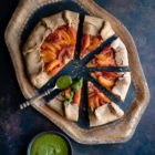 Apricot and Tomato Spelt Galette with Basil Vinaigrette www.thefoodiecorner.gr Photo description: A wooden tray with a galette on it, most of it cut into slices that have been slightly pulled out. Beside one of the slices is a small spoon with basil vinaigrette, some of which has been drizzled on that slice. In the bottom left corner is a small bowl of vinaigrette.