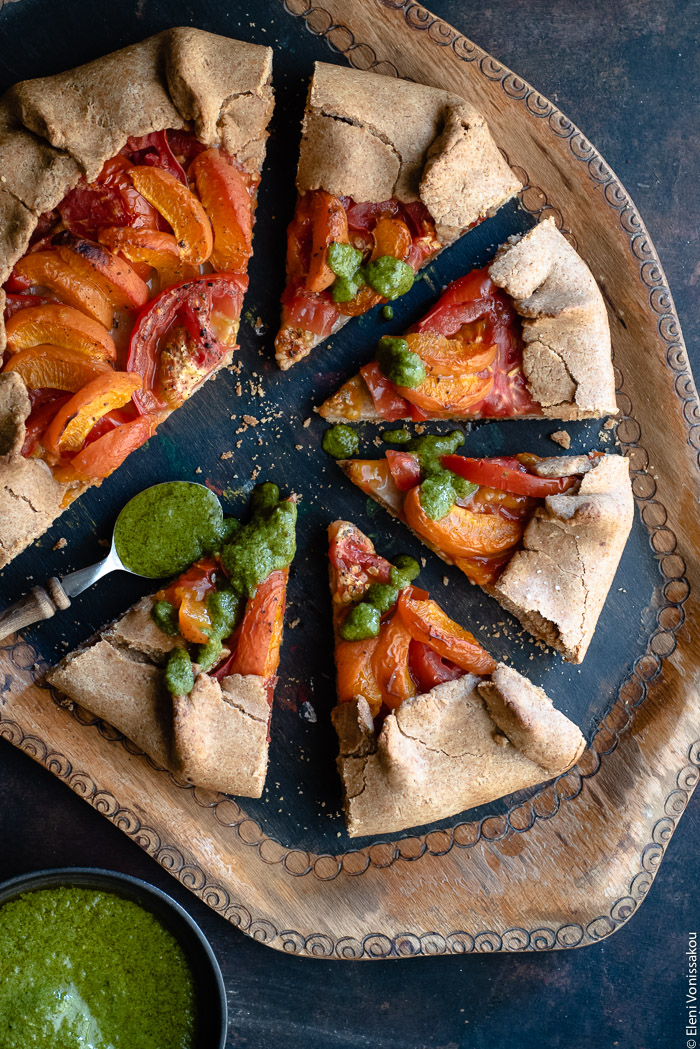 Apricot and Tomato Spelt Galette with Basil Vinaigrette www.thefoodiecorner.gr Photo description: A wooden tray with a galette on it, most of it cut into slices that have been slightly pulled out. Beside one of them is a small spoon with basil vinaigrette, some of which has been drizzled on the slices. In the bottom left corner is a small bowl of vinaigrette, half visible.