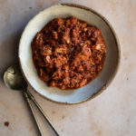 Greek-Style Jackfruit and Tomato Stew (Kokkinisto) in the Slow Cooker www.thefoodiecorner.gr Photo description: A ceramic deep dish full of shredded jackfruit in a red tomato sauce. To the left of the dish are two large serving spoons.