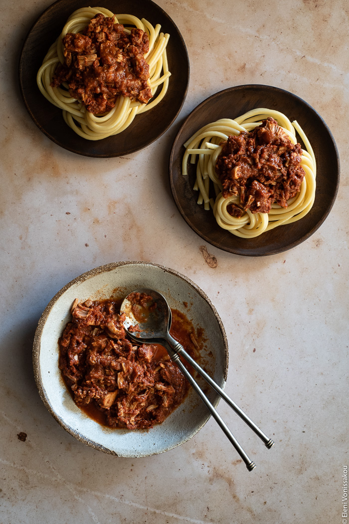 Greek-Style Jackfruit and Tomato Stew (Kokkinisto) in the Slow Cooker www.thefoodiecorner.gr Photo description: A ceramic dish with shredded jackfruit in a tomato sauce, two serving spoons lying inside the dish. To the top of the image are two wooden plates each with a serving of bucatini pasta and jackfruit sauce on top.