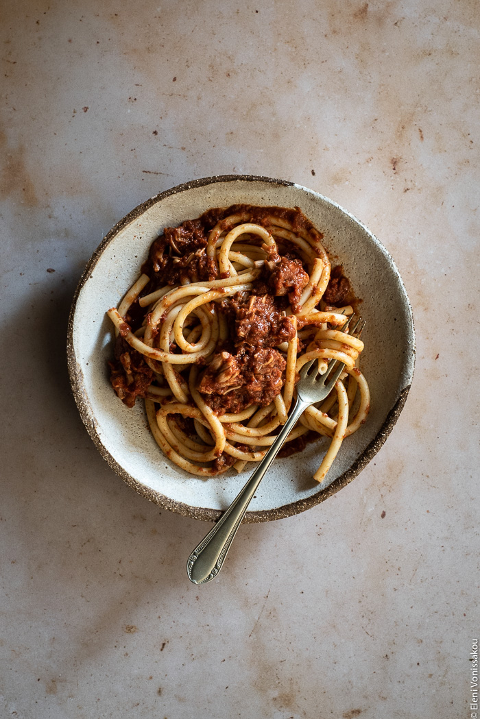 Greek-Style Jackfruit and Tomato Stew (Kokkinisto) in the Slow Cooker www.thefoodiecorner.gr Photo description: A ceramic dish with a large serving of bucatini pasta mixed with shredded jackfruit in tomato sauce. A fork is lying in the pasta. The dish is on a marble surface.