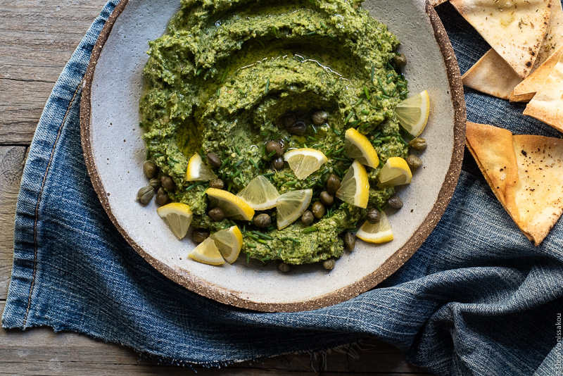 Ideas for Dips with Leftover Herbs www.thefoodiecorner.gr Photo description: A ceramic dish with a vibrant Greek parsley dip garnished with small wedges of lemon, capers and chopped chives. The dish is sitting on a piece of denim and is surrounded by triangular homemade pitta chips.