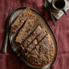 Easy Slow Cooker Olive Oil Banana Bread (Plant Based) www.thefoodiecorner.gr Photo description: A grey ceramic platter with an oval shaped banana bread on it, half of which is sliced, the slices spaced slightly apart. Two slices are lying on their side so we can see the inside of the cake. To the left of the platter is a large vintage knife and to the right is a small ceramic coffee cup and saucer with some coffee in it. A small spoon lies across the saucer. Everything is placed on a creased linen tablecloth.