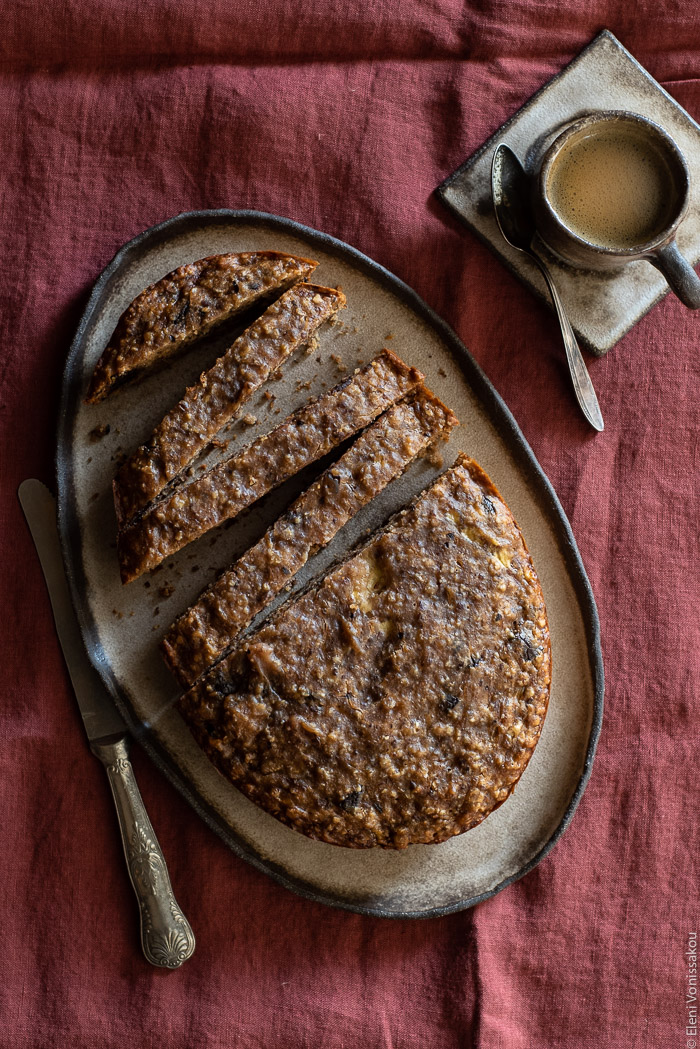 Easy Slow Cooker Olive Oil Banana Bread (Plant Based) www.thefoodiecorner.gr Photo description: A grey ceramic platter with an oval shaped banana bread on it, half of which is sliced, the slices spaced slightly apart. To the left of the platter is a large vintage knife and to the right is a small ceramic coffee cup and saucer with some coffee in it. A small spoon lies across the saucer. Everything is placed on a creased linen tablecloth.