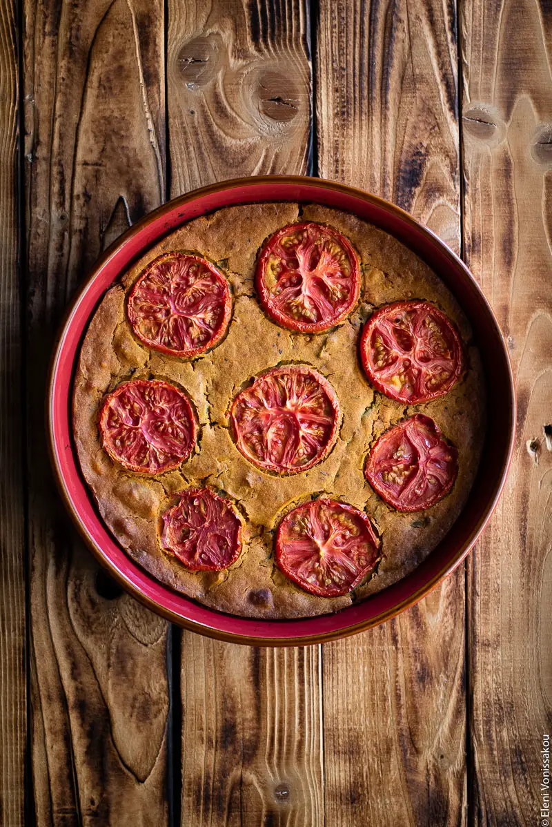 Chickpea Flour Pie (aka Plant-Based Frittata) with Leftover Green Bean Stew www.thefoodiecorner.gr Photo description: A round chickpea flour pie in a red earthenware baking dish on a wooden surface with dark burnt marks. The surface of the pie is deep golden, cracked from cooking, and decorated with cooked slices of tomato.