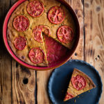 Chickpea Flour Pie (aka Plant-Based Frittata) with Leftover Green Bean Stew www.thefoodiecorner.gr Photo description: A round chickpea flour pie in a red earthenware baking dish on a wooden surface with dark burnt marks. Next to the dish is a black ceramic plate with a piece of pie. The surface of the pie is deep golden, cracked from cooking, and decorated with cooked slices of tomato.