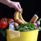 Food Waste and Climate Change: How is the planet affected www.thefoodiecorner.gr Photo description: a metal yellow rubbish bin overflowing with food items such as lettuce, apple, potatoes, tomatoes, cheese and a bottle of milk. A hand is leaving a half visible bunch of bananas in the bin.